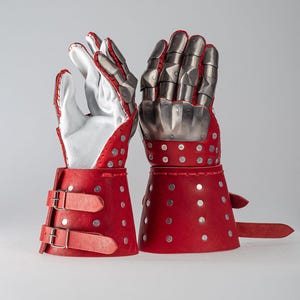 May include: Pair of red and silver medieval gauntlets. The gauntlets feature red leather cuffs with silver rivets and buckles. The hand portion is made of silver metal plates, with white leather gloves inside.