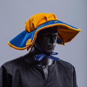 K&ouml;nnte beinhalten: Ein breitkrempiger Hut mit einer gelben Krone und einem blauen Rand. Der Hut hat einen Knopf an der Seite und ein blaues Band, das unter dem Kinn gebunden ist.