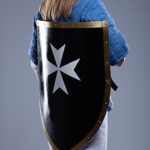 Puede incluir: Escudo negro con un diseño de cruz blanca y borde dorado. El escudo lo sostiene una persona que lleva una chaqueta acolchada azul y pantalones blancos. Un artículo medieval o histórico.