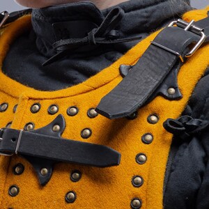 Pode incluir: Close-up de um gambeson medieval amarelo mostarda e preto. A armadura acolchoada apresenta rebites de lat&atilde;o, correias de couro preto com fivelas prateadas e uma gola preta. O logotipo Steel Mastery &eacute; vis&iacute;vel na parte inferior.