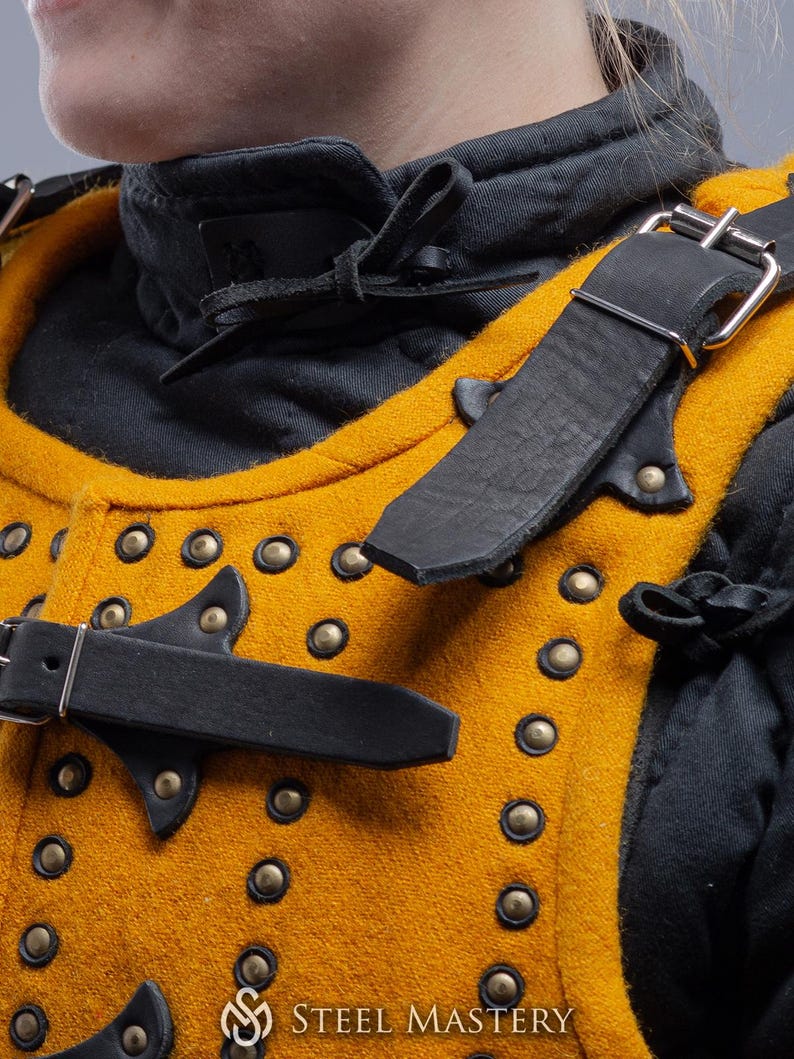 Pode incluir: Close-up de um gambeson medieval amarelo mostarda e preto. A armadura acolchoada apresenta rebites de lat&atilde;o, correias de couro preto com fivelas prateadas e uma gola preta. O logotipo Steel Mastery &eacute; vis&iacute;vel na parte inferior.
