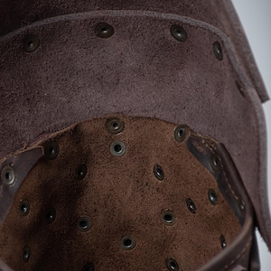 May include: Close-up of the inside of a brown leather helmet. The helmet is lined with brown suede and has numerous small metal rivets. The text "STEEL MASTERY" is visible on the bottom of the helmet.