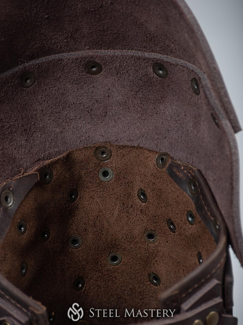 May include: Close-up of the inside of a brown leather helmet. The helmet is lined with brown suede and has numerous small metal rivets. The text "STEEL MASTERY" is visible on the bottom of the helmet.