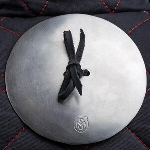 May include: A close-up of a round, silver metal plate with a black leather strap tied in a knot. The plate has a small, embossed logo in the centre. The plate is attached to a black quilted fabric background.