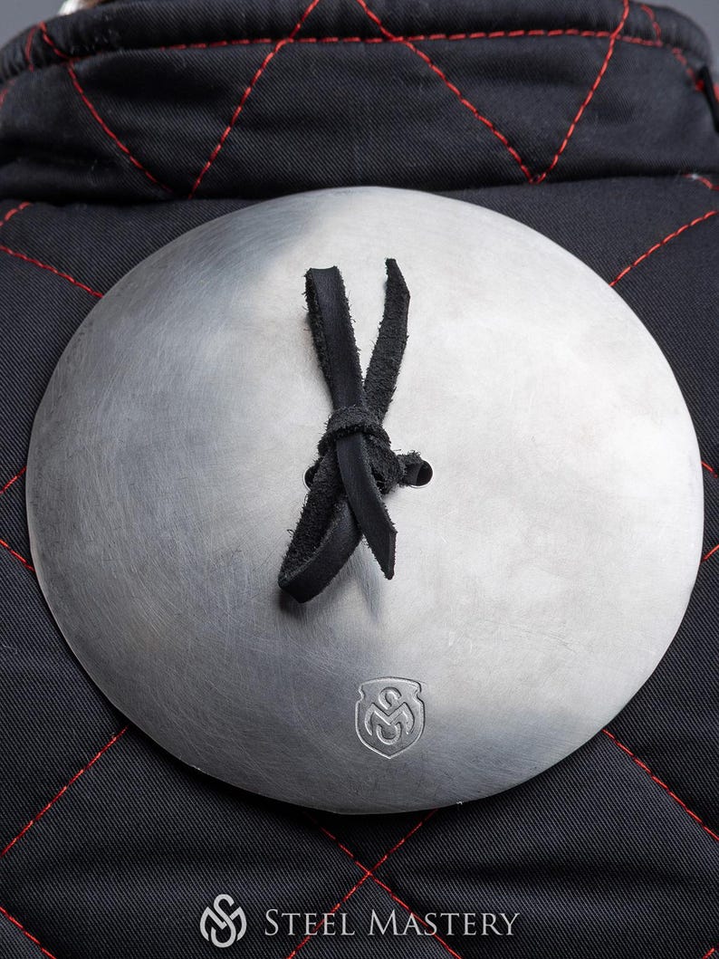May include: A close-up of a round, silver metal plate with a black leather strap tied in a knot. The plate has a small, embossed logo in the centre. The plate is attached to a black quilted fabric background.