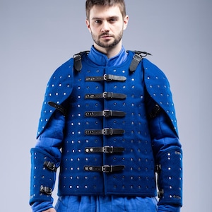 May include: A blue padded gambeson with black leather straps and buckles. The medieval garment features silver rivets and is worn over a blue tunic. The Steel Mastery logo is visible at the bottom.
