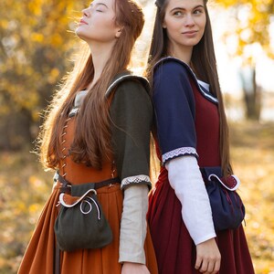 May include: Two women wearing medieval-style dresses stand back-to-back in a forest setting. The woman on the left is wearing a brown dress with a green pouch and a white lace trim. The woman on the right is wearing a red dress with a blue bodice and white lace trim. Both women have long brown hair.