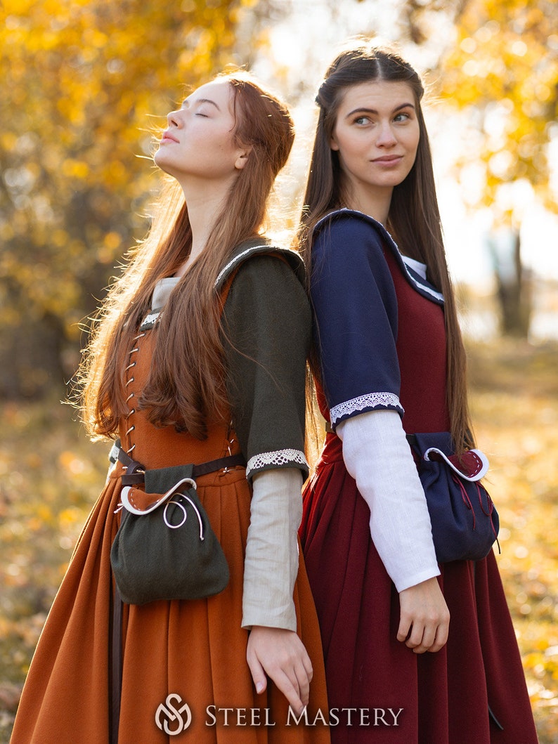 May include: Two women wearing medieval-style dresses stand back-to-back in a forest setting. The woman on the left is wearing a brown dress with a green pouch and a white lace trim. The woman on the right is wearing a red dress with a blue bodice and white lace trim. Both women have long brown hair.