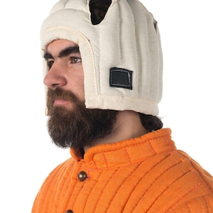 Puede incluir: Un casco acolchado blanco con un parche negro en el lateral. El casco lo lleva una persona con barba y una chaqueta acolchada naranja.