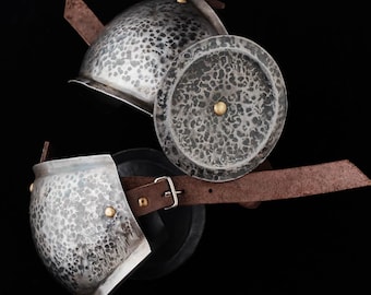 May include: Two pieces of medieval armor, likely for the arm, are displayed against a black background. The metal pieces have a hammered texture and are connected by brown leather straps with buckles. The armor is silver with gold rivets.