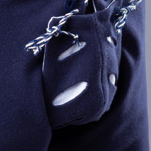 Op de afbeelding: Een close-up van een marineblauw kledingstuk met een witte zak met een decoratief vetersluiting. De zak heeft drie ovale uitsparingen. Het kledingstuk heeft een wit en marineblauw koord dat langs de zijkant loopt.