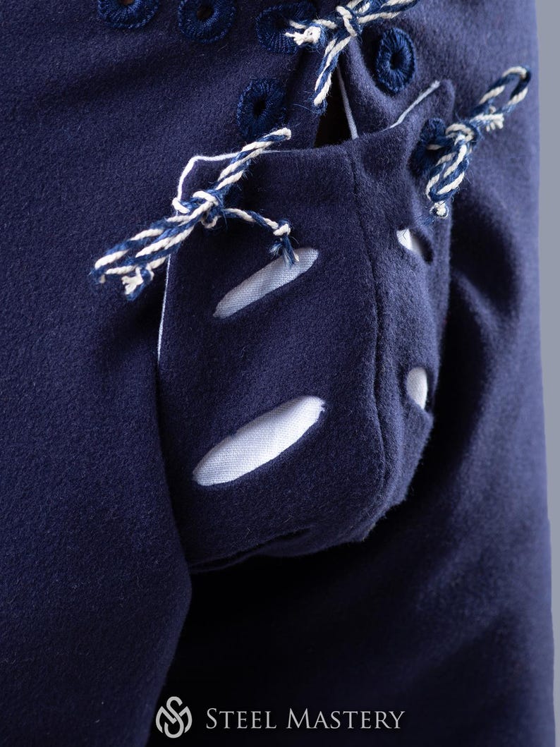 Op de afbeelding: Een close-up van een marineblauw kledingstuk met een witte zak met een decoratief vetersluiting. De zak heeft drie ovale uitsparingen. Het kledingstuk heeft een wit en marineblauw koord dat langs de zijkant loopt.