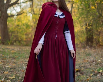 May include: A woman wearing a long, burgundy dress with a matching hooded cape. The dress has a lace-up bodice and the cape has a long train. She is standing in a forest setting with fallen leaves on the ground.