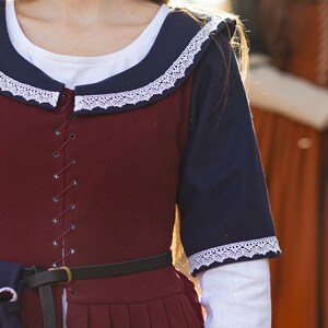 May include: A burgundy dress with a navy blue collar and sleeves. The dress has a white lace trim and a lace-up front. The dress is cinched at the waist with a black leather belt.