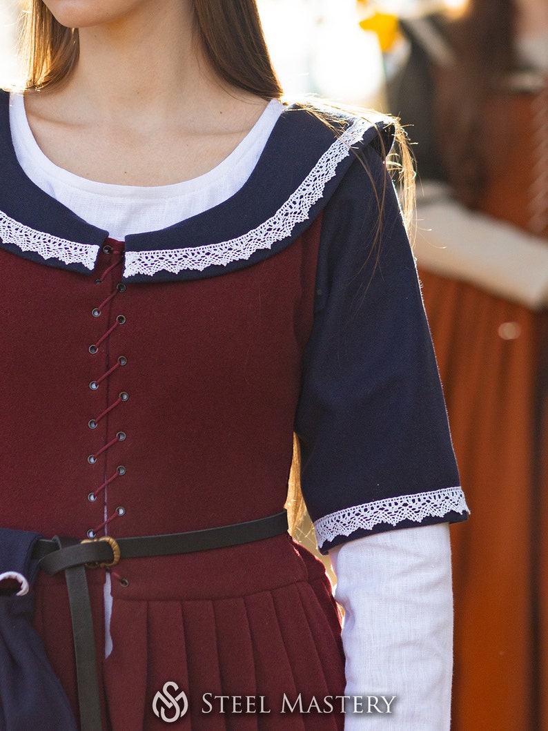 May include: A burgundy dress with a navy blue collar and sleeves. The dress has a white lace trim and a lace-up front. The dress is cinched at the waist with a black leather belt.