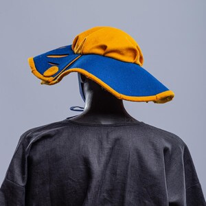 K&ouml;nnte beinhalten: Ein Hut mit breiter Krempe, blauer Krone und gelbem Rand. Der Hut hat ein einzigartiges Design mit Ausschnitten in der Krempe.