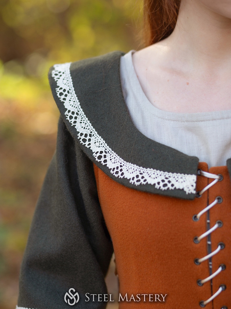 May include: A close-up of a brown and green medieval-style dress with a white lace trim around the neckline. The dress has a laced-up front and a fitted bodice.