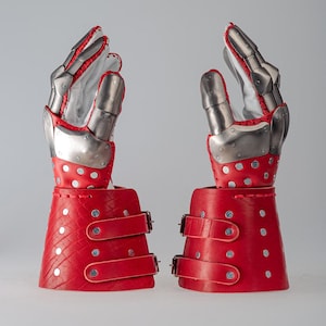 May include: Pair of medieval-style gauntlets. The gauntlets feature articulated metal hand armor with red leather accents and wrist cuffs. The red leather cuffs have silver rivets and adjustable leather straps with buckles.