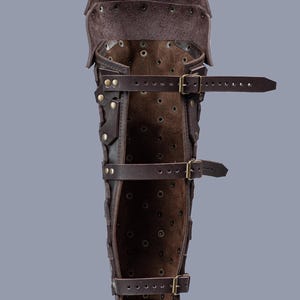 May include: Brown leather leg armor with buckles and straps. The armor is made of multiple pieces of leather that are stitched together. The armor is designed to protect the shin and calf.
