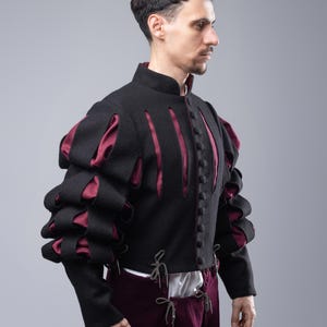 May include: A black doublet with burgundy accents and puffed sleeves. The doublet has a stand-up collar and a button-up front. The sleeves are gathered at the shoulders and have a series of vertical cuts that reveal the burgundy lining. The doublet is worn over a white shirt and burgundy pants.