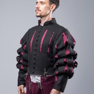May include: A black doublet with burgundy accents and puffed sleeves. The doublet has a high collar and is fastened with buttons. The sleeves are gathered at the elbow and wrist. The doublet is worn over a white shirt and burgundy pants.