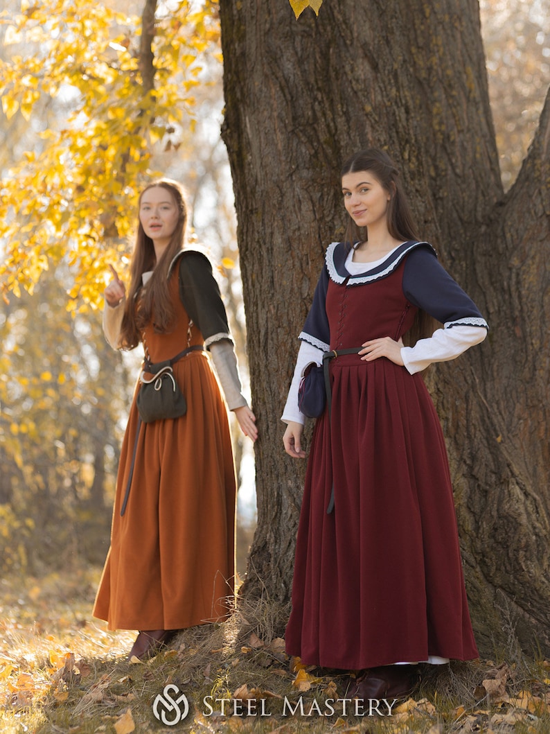 May include: Two women wearing medieval-style dresses stand in a forest. The woman on the left is wearing a brown dress with a black pouch. The woman on the right is wearing a red dress with a blue and white collar and a black pouch.