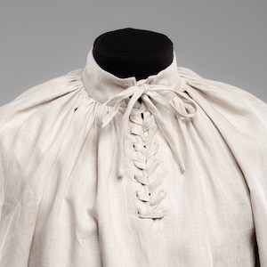 May include: A beige linen shirt with a lace-up neckline. The shirt has a gathered, puffed look around the neckline.