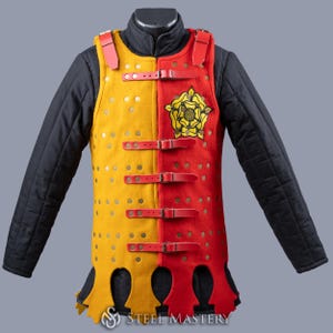 Puede incluir: Un chaleco medieval de gambesón de dos tonos en amarillo y rojo, asegurado con correas y hebillas de cuero rojo. El lado amarillo tiene numerosos remaches metálicos. El lado rojo presenta un emblema floral dorado. El chaleco se usa sobre una prenda interior acolchada negra.