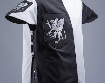 Puede incluir: Una túnica medieval en blanco y negro con un emblema de dragón en el pecho y las mangas. La túnica tiene una abertura frontal con botones y un dobladillo decorativo. Las mangas son cortas y la túnica se exhibe en un maniquí.
