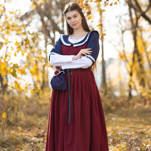 May include: A woman wearing a long, burgundy dress with a white blouse underneath. She is wearing a blue shawl with white trim and a black belt. She is standing in a forest with fall foliage.