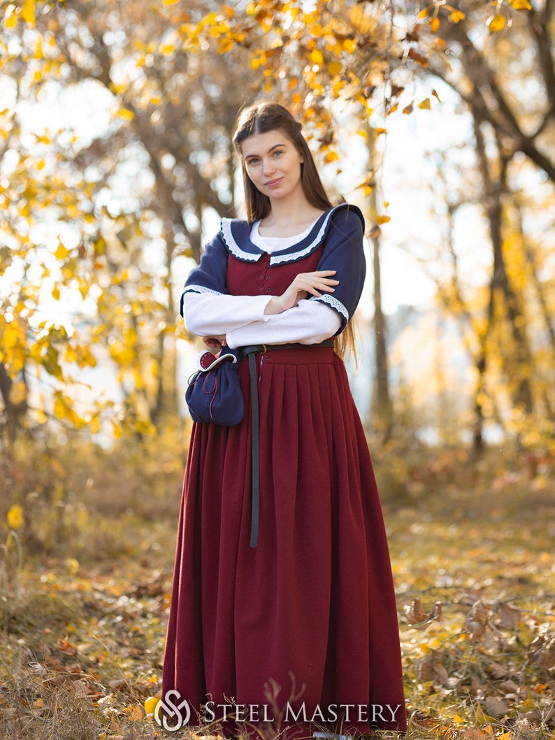May include: A woman wearing a long, burgundy dress with a white blouse underneath. She is wearing a blue shawl with white trim and a black belt. She is standing in a forest with fall foliage.