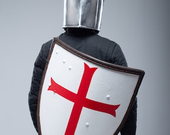 May include: A person wearing a medieval helmet and chainmail holds a large, white shield with a red cross. The shield is decorated with small metal rivets.