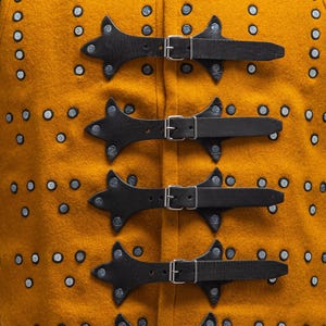 Pode incluir: Close-up de um colete medieval amarelo mostarda com tiras de couro preto e fivelas prateadas. O colete tem rebites decorativos prateados e o logotipo "STEEL MASTERY" na parte inferior.