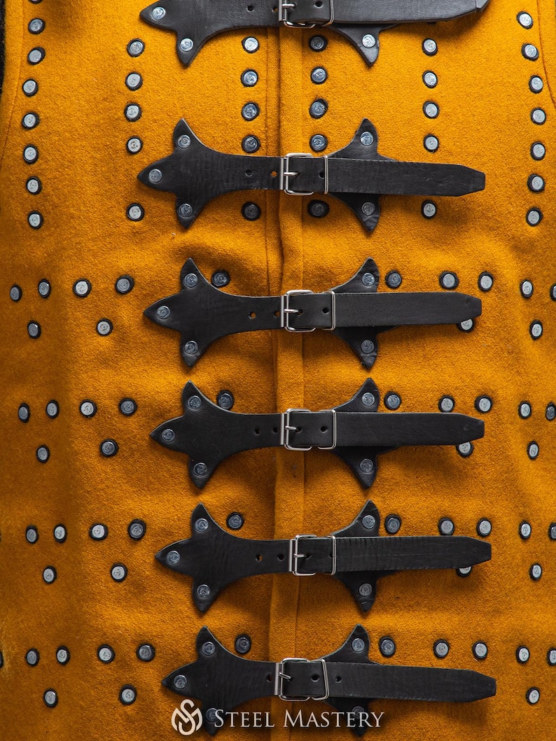 Pode incluir: Close-up de um colete medieval amarelo mostarda com tiras de couro preto e fivelas prateadas. O colete tem rebites decorativos prateados e o logotipo "STEEL MASTERY" na parte inferior.