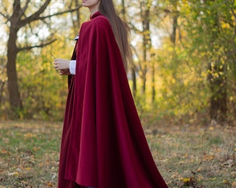 May include: A long, flowing burgundy velvet cloak with a deep blue trim. The cloak is worn over a white collared shirt and a dark dress. The image is set in a forest with autumn foliage.