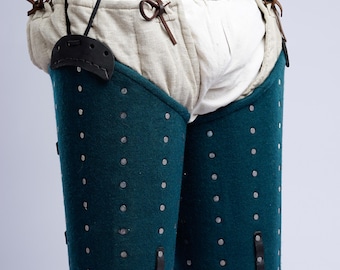 May include: Medieval teal leg armor with rivets and leather straps. The armor is worn over padded fabric and secured with a linen belt and leather straps. The image shows a close-up of the armor, highlighting its construction and design.