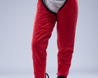 May include: Red padded pants with vertical stitching, worn with a white undergarment and a red belt. The pants are part of a historical costume, paired with brown leather boots. The Steel Mastery logo is visible at the bottom.