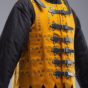 Pode incluir: Gambeson medieval amarelo mostarda com tiras e fivelas de couro preto. O colete &eacute; cravejado com rebites prateados e tem uma bainha pontiaguda. A pe&ccedil;a &eacute; usada sobre uma roupa interior acolchoada preta. O logotipo Steel Mastery &eacute; vis&iacute;vel na parte inferior.