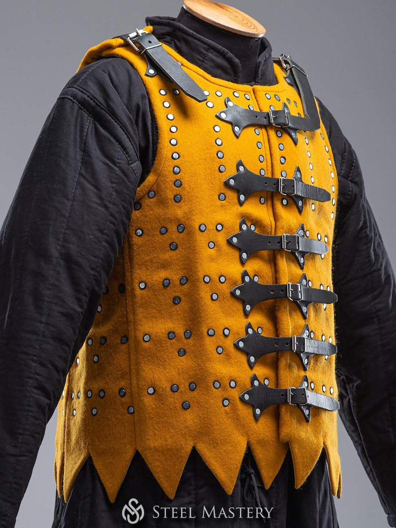 Pode incluir: Gambeson medieval amarelo mostarda com tiras e fivelas de couro preto. O colete &eacute; cravejado com rebites prateados e tem uma bainha pontiaguda. A pe&ccedil;a &eacute; usada sobre uma roupa interior acolchoada preta. O logotipo Steel Mastery &eacute; vis&iacute;vel na parte inferior.