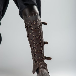 May include: Brown leather leg armor with a hexagonal pattern and buckles. The armor is worn over a pair of brown leather boots.
