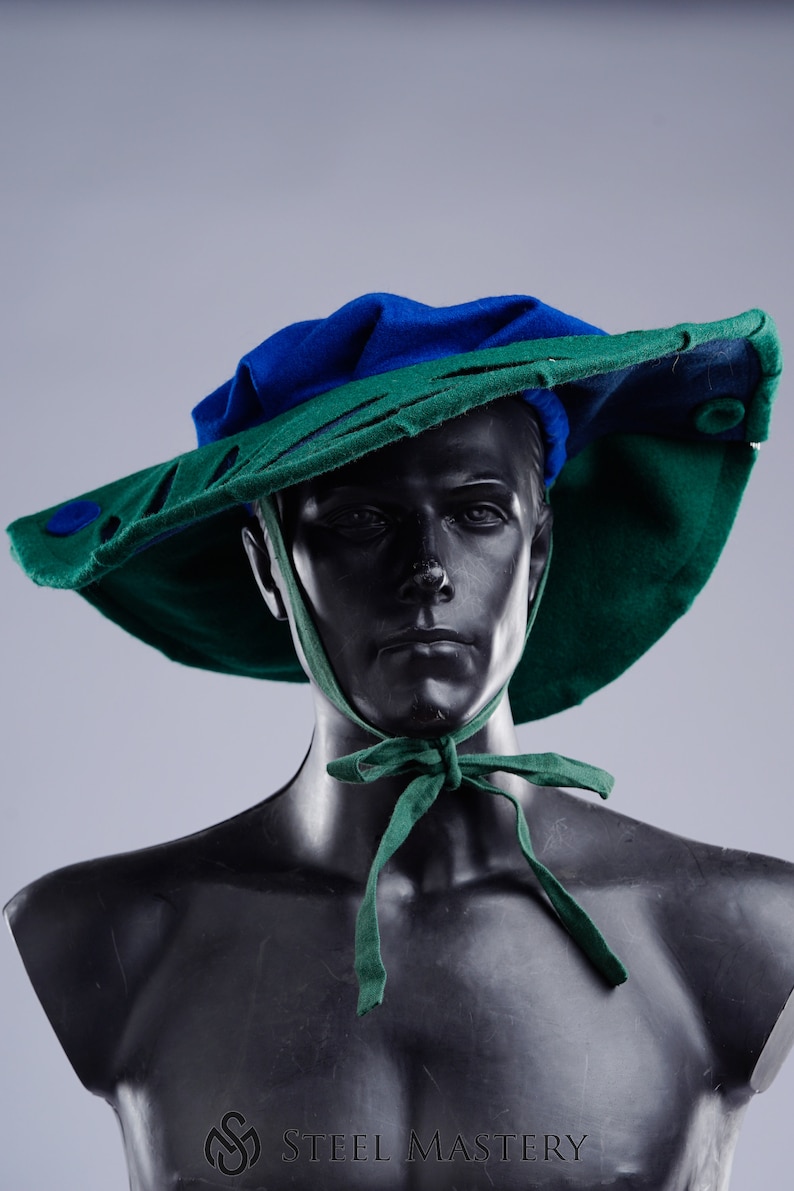 K&ouml;nnte beinhalten: Ein gr&uuml;ner und blauer Filzhut mit breiter Krempe und einem ausgeschnittenen Design. Der Hut ist mit einem gr&uuml;nen Band unter dem Kinn gebunden.