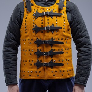 Pode incluir: Colete medieval amarelo mostarda com tiras e fivelas de couro preto. O colete &eacute; cravejado com pequenos rebites e apresenta uma gola alta. O logotipo Steel Mastery &eacute; vis&iacute;vel na parte inferior.