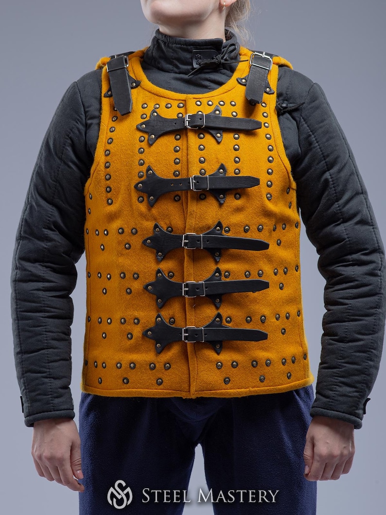 Pode incluir: Colete medieval amarelo mostarda com tiras e fivelas de couro preto. O colete &eacute; cravejado com pequenos rebites e apresenta uma gola alta. O logotipo Steel Mastery &eacute; vis&iacute;vel na parte inferior.