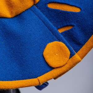 K&ouml;nnte beinhalten: Nahaufnahme eines blauen und goldenen Stoffes mit einem runden, goldenen Knopf. Der Stoff hat eine strukturierte Oberfl&auml;che und einen kontrastierenden Besatz.