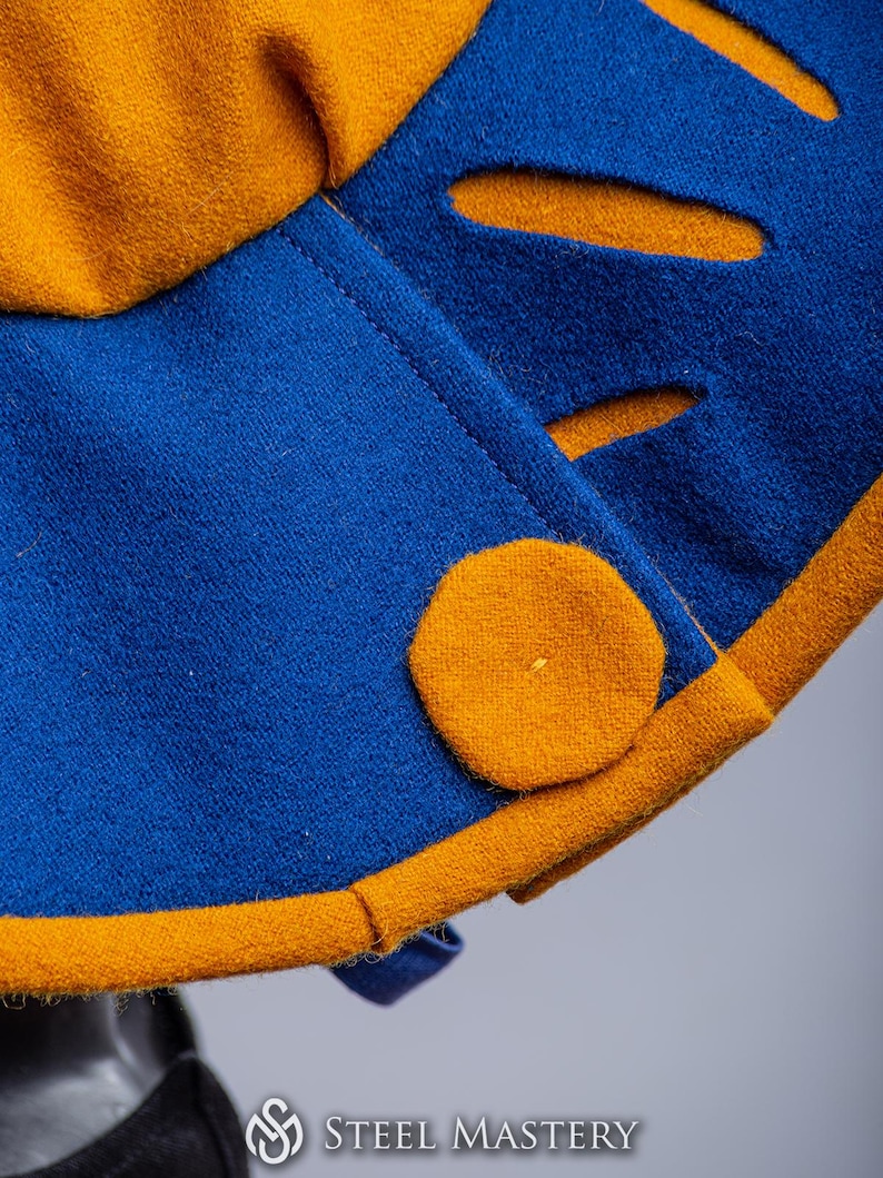K&ouml;nnte beinhalten: Nahaufnahme eines blauen und goldenen Stoffes mit einem runden, goldenen Knopf. Der Stoff hat eine strukturierte Oberfl&auml;che und einen kontrastierenden Besatz.