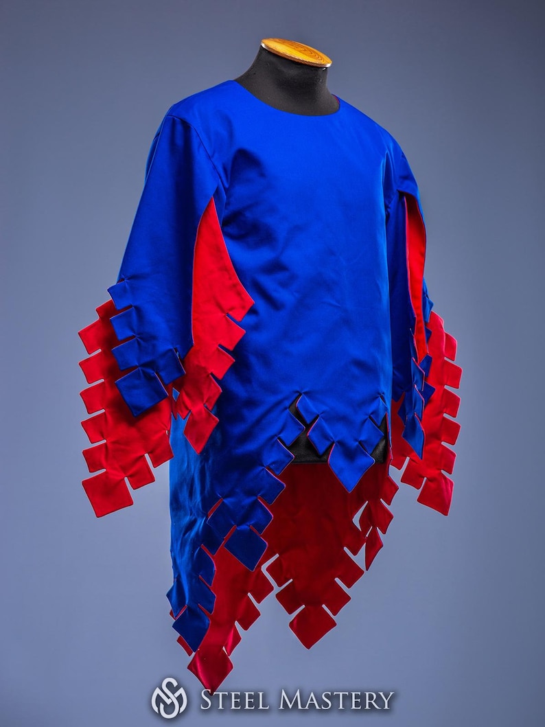 May include: A blue tunic with red detailing, presented on a mannequin. The garment has a round neckline and long sleeves. Red, square cutouts adorn the sleeves and hem. The Steel Mastery logo is visible at the bottom of the image.