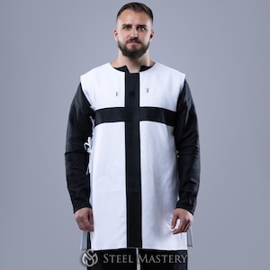 May include: A white tunic with a large black cross design on the front. The long sleeves are black, and the tunic has a high neckline. The bottom of the tunic is visible, and the Steel Mastery logo is at the bottom.