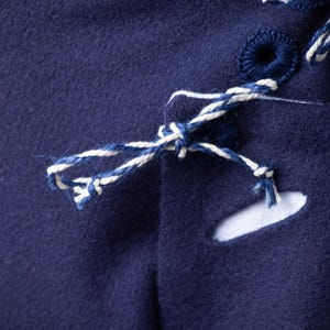 Op de afbeelding: Close-up van een marineblauwe stof met witte en blauwe stiksels. De stof heeft een aantal kleine gaten en een wit koord dat door de gaten is gebonden.