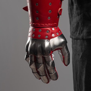 May include: A red leather gauntlet with silver metal plates covering the hand and fingers. The gauntlet is studded with silver rivets and features a red leather cuff. The design suggests medieval armor.