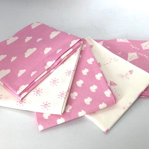 May include: Folded fabric squares in pink and cream, patterned with clouds, suns, and kites. The pink fabric displays white clouds and kites. The cream fabric features sun and kite designs. Suitable for sewing or craft projects, in metric measurements.